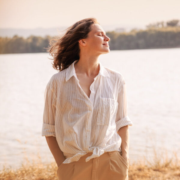 Жена, стояща навън край езеро, се наслаждава на слънцето със затворени очи